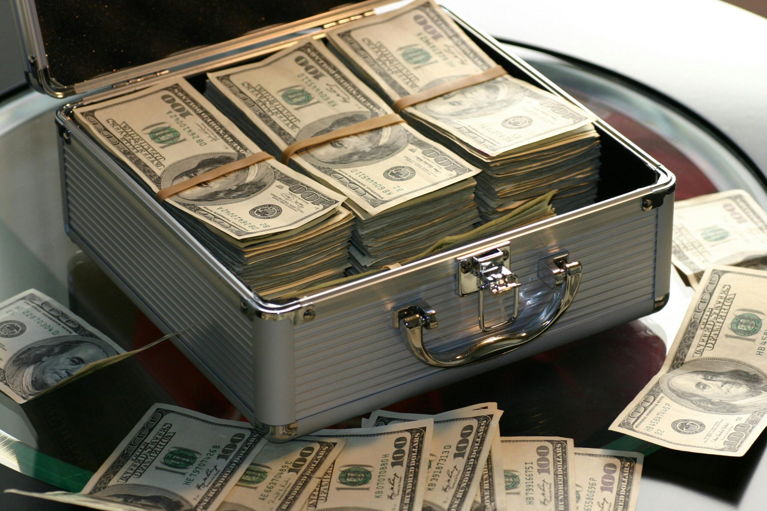 Open briefcase filled with stacks of hundred dollar bills on a glass table, representing wealth.