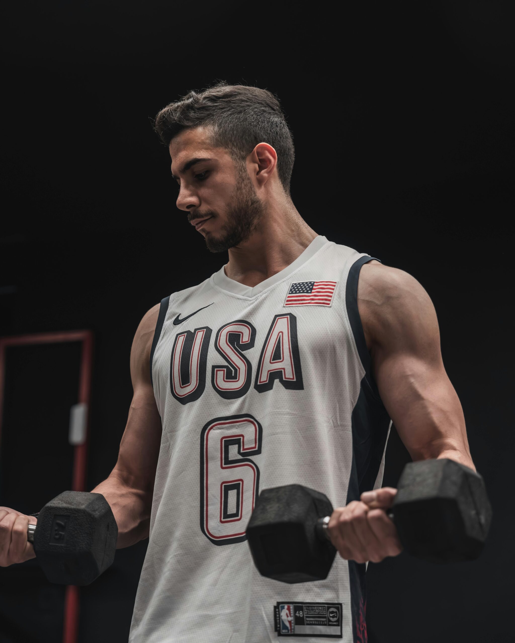 Young male athlete in USA jersey lifting dumbbells indoors, showcasing strength and fitness.