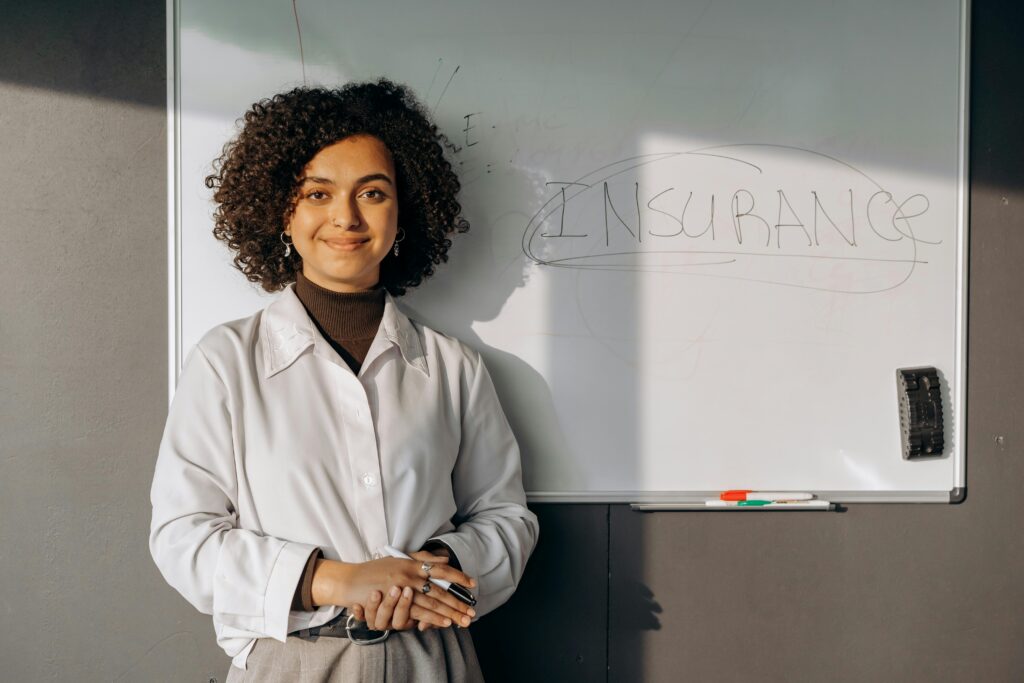 Smiling woman with curly hair standing by a whiteboard with 'insurance' written on it, conveying professionalism.