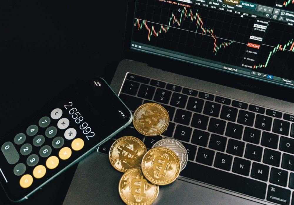 A still life of Bitcoin coins, a smartphone calculator, and a trading chart on a laptop.